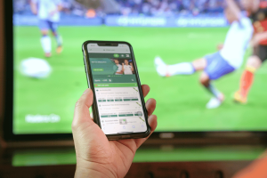 The study warns that gambling ads are driving soccer betting during the 2022 World Cup despite UK safeguards. Man holding a smartphone showing a sports betting app while watching a live soccer match on television in the background.