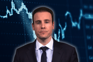 The curious case of Michael Selig and his predictions about markets. CFTC chair Mike Selig in a dark suit and tie faces the camera against a blue background featuring stylized financial charts and market data graphics.