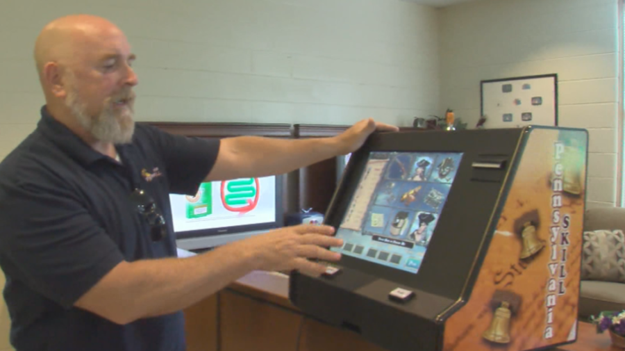 Rick Goodling é um homem careca e barbudo, vestindo uma camisa pólo escura, demonstrando uma máquina de jogos de vídeo ou de habilidade dentro de um escritório.