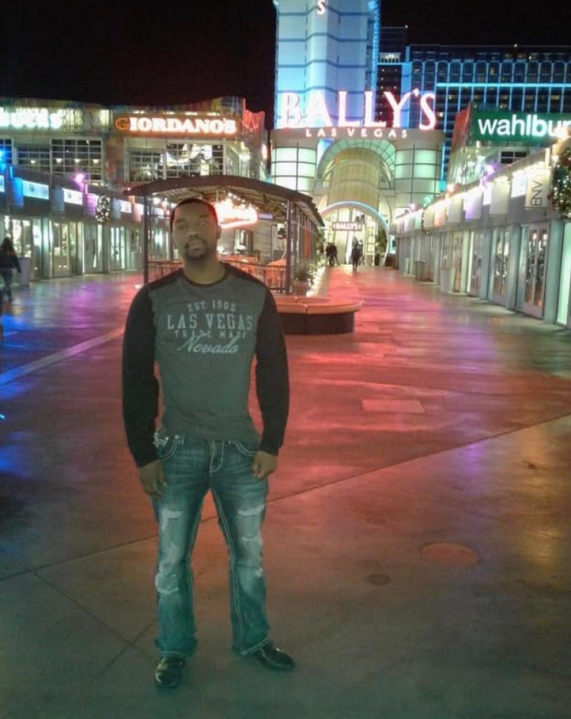 Shaun Joseph Benward está em um calçadão ao ar livre iluminado à noite em frente ao Bally's Las Vegas, com placas de Giordano's e Wahlburgers brilhando ao fundo.