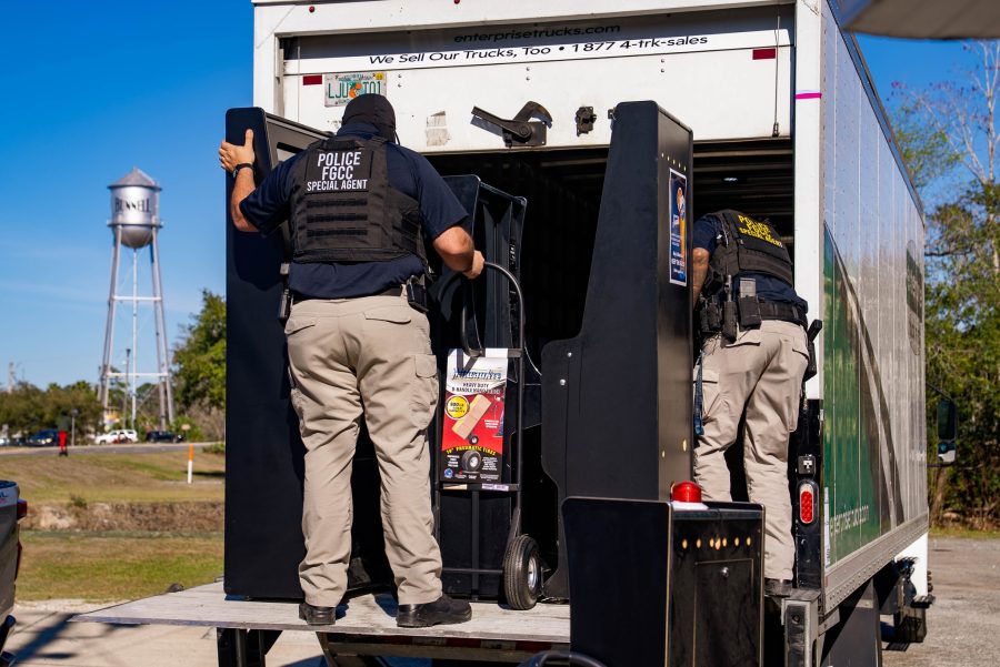 Dois policiais usando coletes táticos rotulados como “Agente Especial da Polícia FGCC” carregam grandes máquinas de jogos pretas na traseira de um caminhão durante uma operação diurna.