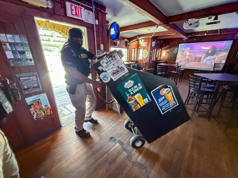 Um policial conduz uma grande máquina de jogos em um carrinho pelo interior de um bar, passando por paredes de madeira, mesas altas e uma tela de televisão.