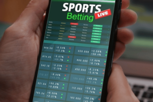 ACMA says betting firms' failures undermine BetStop national self-exclusion system. Hand holding a smartphone displaying a sports betting app interface with live odds, match listings, and fluctuating betting data.