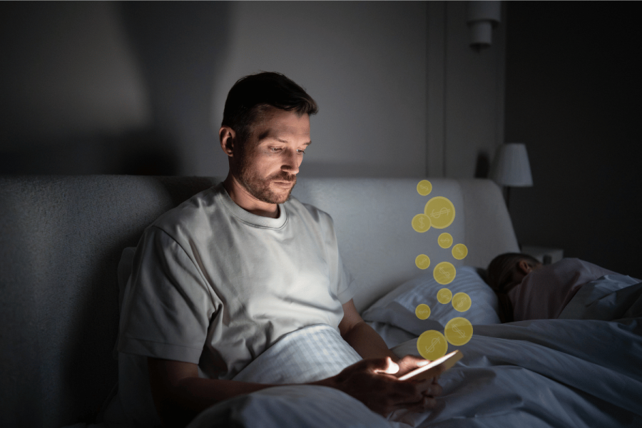 Man lying in bed at night looking at a glowing smartphone while stylized gold coin icons float upward from the screen, suggesting online gambling or betting activity as another person sleeps beside him in a dimly lit bedroom. Digital betting boom reshapes American gambling habits raising financial harm concerns