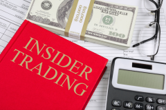From Trading Places to war markets how insider trading slipped into prediction platforms. A red book labeled “Insider Trading” lies on paperwork beside a calculator, eyeglasses, and a stack of U.S. hundred-dollar bills.