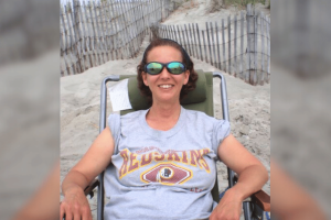 Luzerne church worker sentenced to prison after stealing funds for gambling. Woman wearing sunglasses relaxes in a beach chair on a sandy dune path, smiling while wearing a gray Washington Redskins T-shirt.