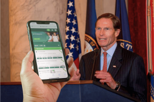 U.S. Senator speaking at podium with American flags while smartphone displays sports betting app interface in foreground, illustrating concerns over gambling partnerships and sports integrity. Blumenthal presses sports leagues over betting ties and new integrity concerns
