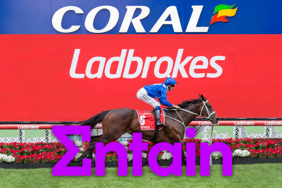 Entain begins restructuring with 500 job cuts as tax pressures mount. Coral and Ladbrokes branding displayed on a racetrack billboard as a jockey rides a racehorse past, with a large Entain logo overlaid in the foreground.