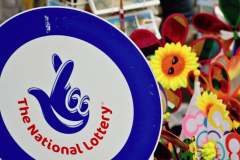A sign featuring "The National Lottery" logo with a blue background and a hand symbol with crossed fingers. In the background, there are colorful pinwheels and sunflowers with smiling faces, likely part of a festive street stall or market. UK's National Lottery to temporarily shut down for major tech updates. UK's National Lottery returns to '90s 'It Could Be You' tagline. Court of Appeal blocks Richard Desmond companies National Lottery license challenge